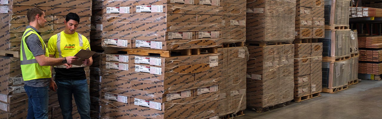 Two employees in warehouse, standing in front of pallets of boxes